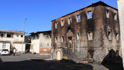 The government will demolish the buildings gutted by fire, including this brick building, left, from the nation's colonial past. (IWN photo)