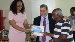 Prime Minister Dr. Ralph Gonsalves, centre, and Natasha Devonish, Manager of Finance at AIA, present a certificate to former property owner, Sam Agard. (iWN photo)