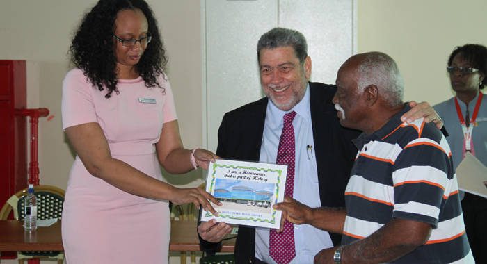Prime Minister Dr. Ralph Gonsalves, centre, and Natasha Devonish, Manager of Finance at AIA, present a certificate to former property owner, Sam Agard. (iWN photo)
